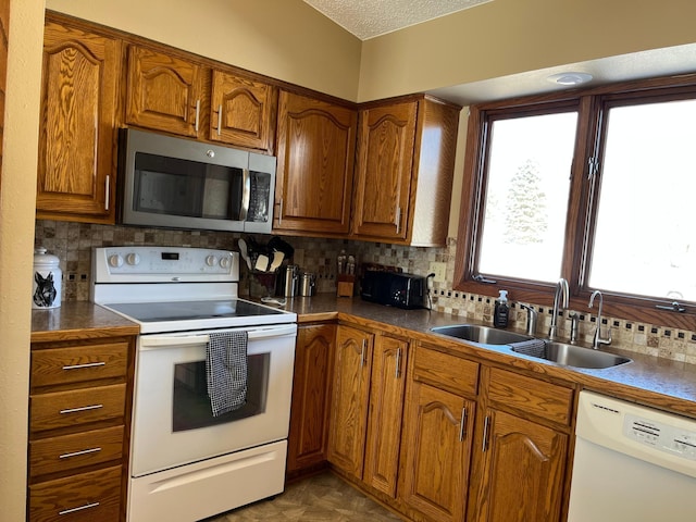 kitchen with brown cabinetry, white appliances, a sink, backsplash, and dark countertops