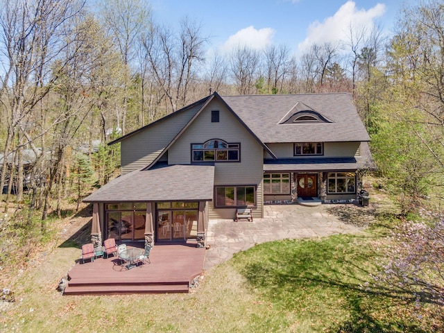 rear view of house featuring a wooden deck, roof with shingles, stone siding, and a patio area