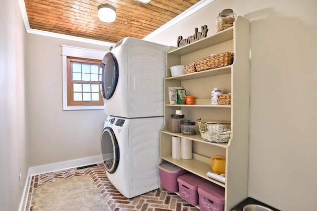clothes washing area featuring stacked washing machine and dryer, baseboards, brick floors, wood ceiling, and ornamental molding