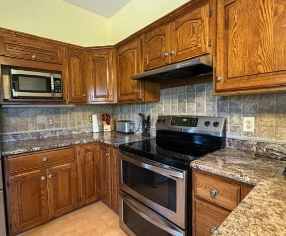 29441 108th St NW, Princeton, MN 55371 - kitchen with appliances with stainless steel finishes, under cabinet range hood, brown cabinets, dark stone countertops, and decorative backsplash