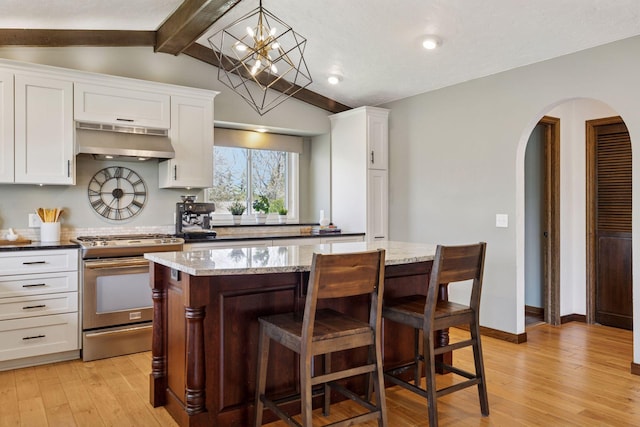 kitchen with stainless steel gas range oven, under cabinet range hood, arched walkways, white cabinets, and light wood-style floors