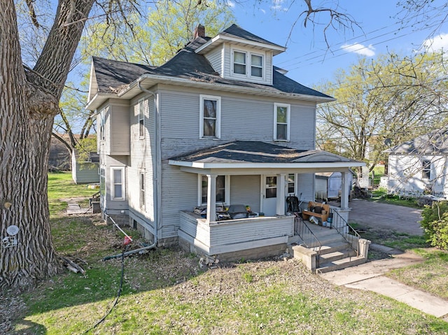 american foursquare style home with a porch, a chimney, a front yard, and a shingled roof