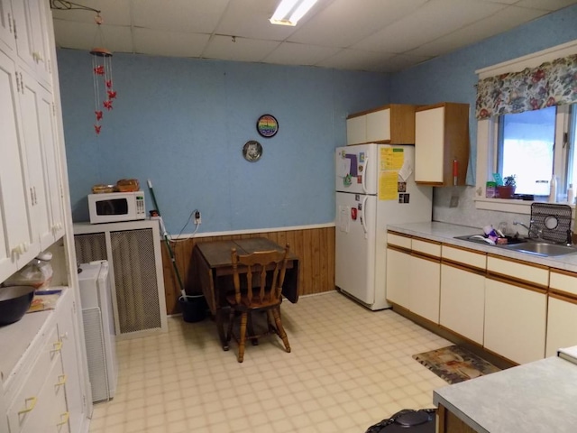 kitchen featuring white appliances, a paneled ceiling, light flooring, a sink, and wainscoting