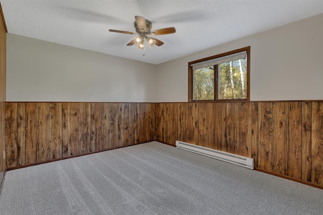 carpeted empty room with a baseboard heating unit, wood walls, ceiling fan, wainscoting, and a textured ceiling