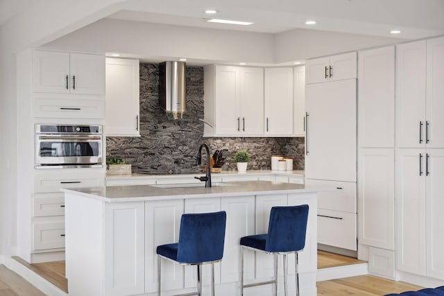 kitchen featuring oven, light countertops, white cabinetry, a sink, and wall chimney range hood