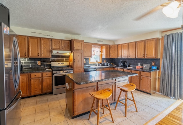 kitchen with appliances with stainless steel finishes, a sink, a center island, brown cabinetry, and a textured ceiling