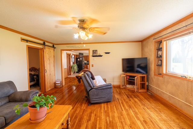 living area with light wood-type flooring, crown molding, ceiling fan, a barn door, and baseboards