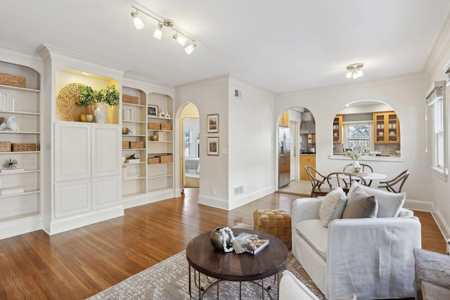 living area with arched walkways, wood finished floors, baseboards, crown molding, and built in shelves