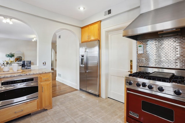 kitchen featuring appliances with stainless steel finishes, wall chimney exhaust hood, arched walkways, backsplash, and light stone countertops