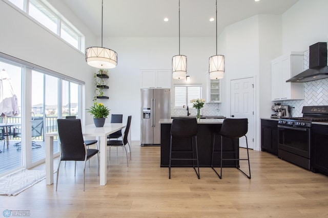 kitchen with black gas range, stainless steel fridge with ice dispenser, wall chimney range hood, a high ceiling, and light countertops