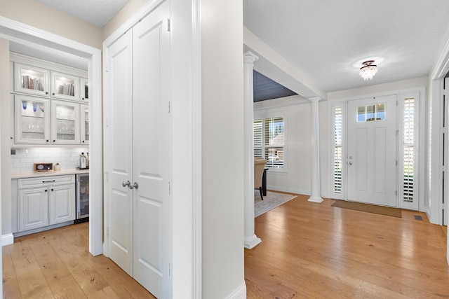 foyer with decorative columns, light wood finished floors, baseboards, wine cooler, and a textured ceiling