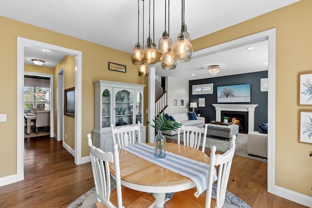 dining area with wood finished floors, stairs, a warm lit fireplace, a chandelier, and baseboards
