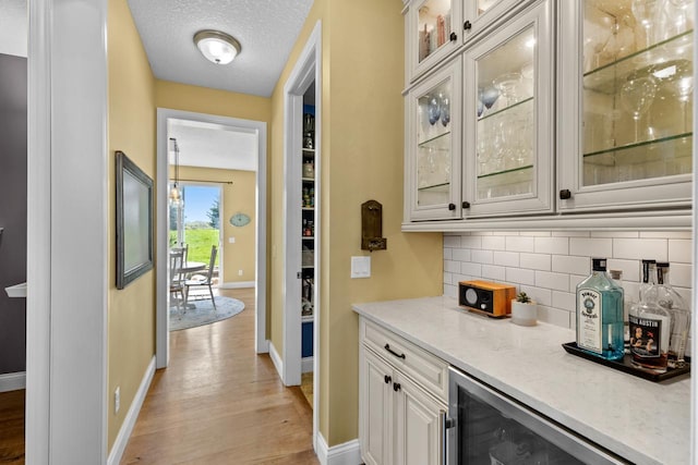 indoor dry bar featuring baseboards, tasteful backsplash, light wood finished floors, wine cooler, and a textured ceiling