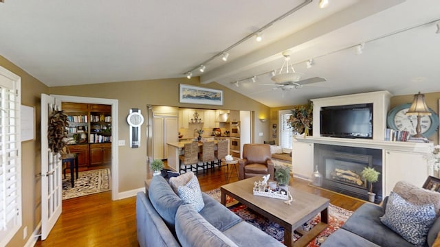 living room with track lighting, wood finished floors, a fireplace with flush hearth, vaulted ceiling, and a ceiling fan