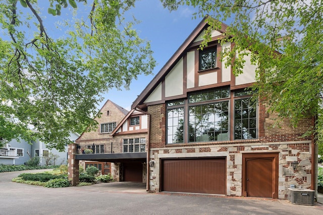 tudor home with stone siding, central air condition unit, stucco siding, a garage, and asphalt driveway