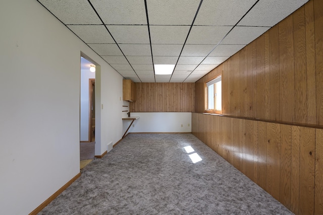 carpeted empty room with wooden walls, a drop ceiling, and baseboards