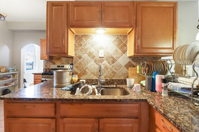 kitchen with a sink, arched walkways, backsplash, and dark stone counters