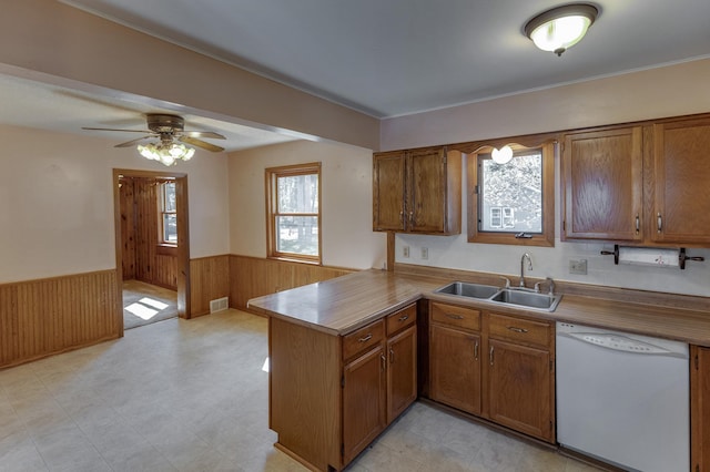 kitchen featuring white dishwasher, a sink, a wainscoted wall, a peninsula, and brown cabinets