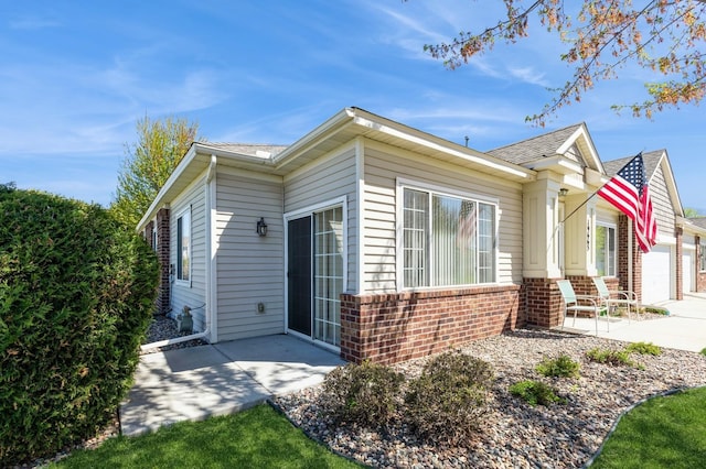view of home's exterior featuring brick siding, a patio area, concrete driveway, roof with shingles, and an attached garage