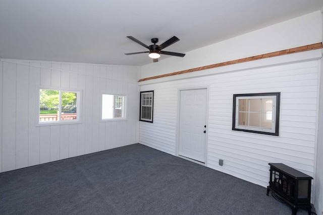 empty room featuring dark carpet, a ceiling fan, a wood stove, and wood walls
