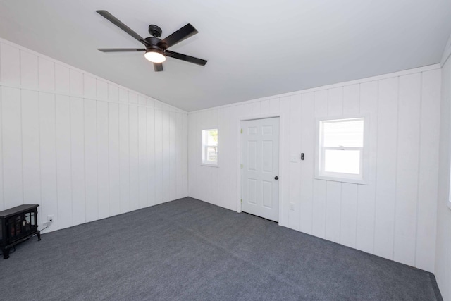 empty room with dark colored carpet, a ceiling fan, lofted ceiling, a wood stove, and wood walls