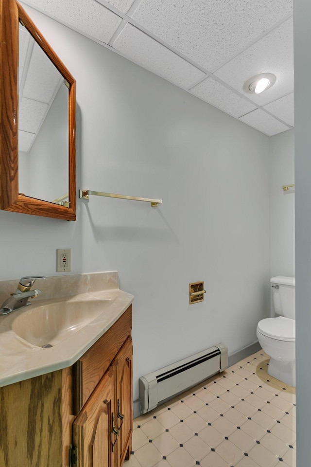 half bathroom with a baseboard radiator, tile patterned floors, toilet, a drop ceiling, and vanity