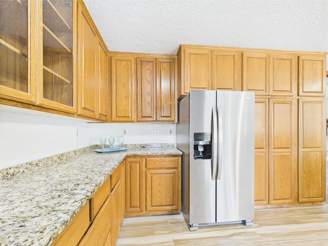 kitchen with stainless steel fridge with ice dispenser, light stone countertops, glass insert cabinets, brown cabinets, and light wood-type flooring