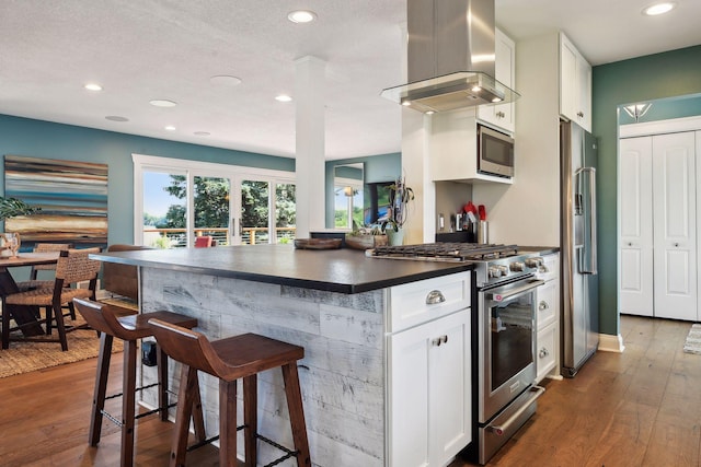 kitchen with appliances with stainless steel finishes, wall chimney range hood, a kitchen bar, dark countertops, and recessed lighting