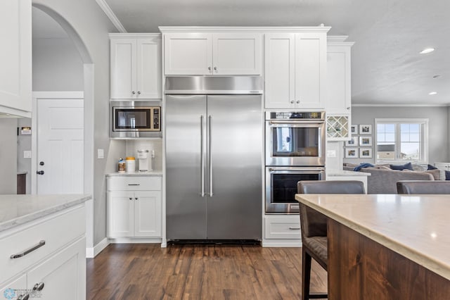 kitchen featuring built in appliances, crown molding, arched walkways, dark wood finished floors, and white cabinets