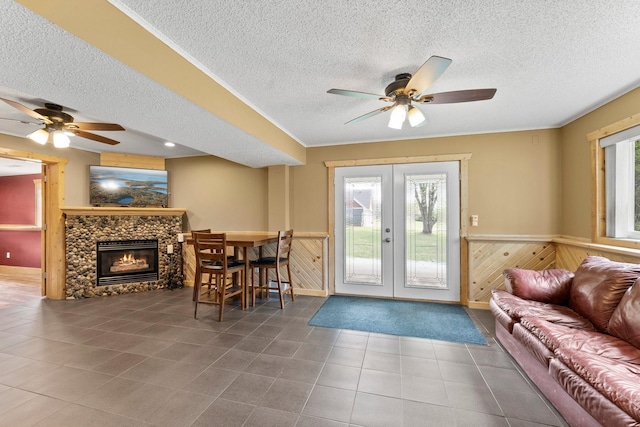 tiled living room featuring french doors, a textured ceiling, ceiling fan, wainscoting, and wood walls