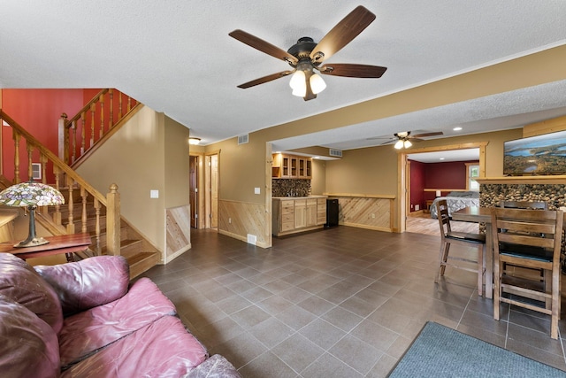 living area with ceiling fan, a wainscoted wall, stairway, a textured ceiling, and dark tile patterned flooring