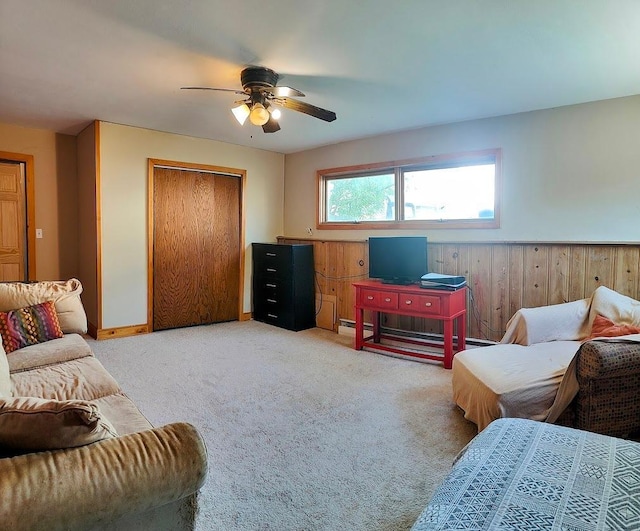 living area featuring a ceiling fan, light colored carpet, a wainscoted wall, a baseboard radiator, and wood walls