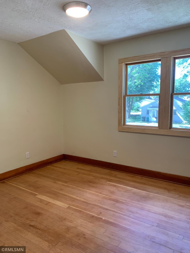 additional living space with a textured ceiling, light wood-type flooring, healthy amount of natural light, baseboards, and vaulted ceiling