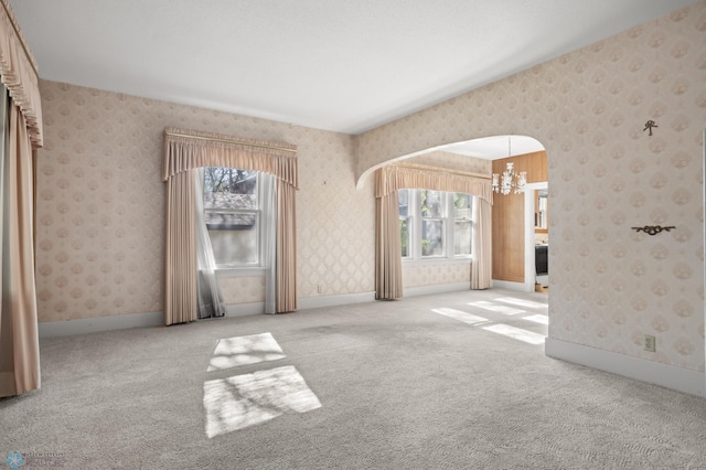 carpeted empty room with a chandelier, arched walkways, baseboards, and wallpapered walls
