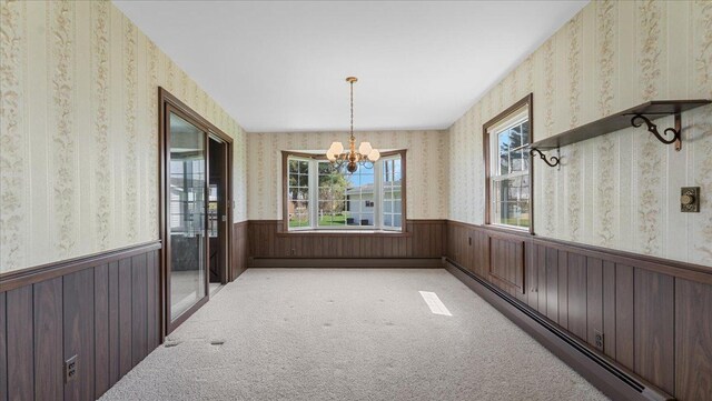 unfurnished dining area with a wainscoted wall, wallpapered walls, baseboard heating, a chandelier, and light colored carpet