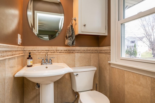 half bathroom with plenty of natural light, a wainscoted wall, toilet, and tile walls