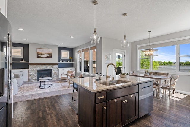 kitchen with a sink, dishwasher, a stone fireplace, dark brown cabinetry, and dark wood finished floors