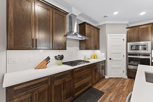 kitchen with appliances with stainless steel finishes, wall chimney range hood, ornamental molding, light countertops, and dark brown cabinets