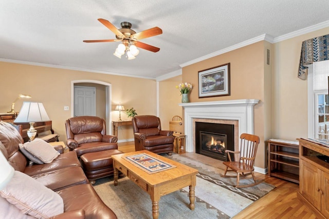 living room with light wood finished floors, a glass covered fireplace, ornamental molding, a ceiling fan, and arched walkways