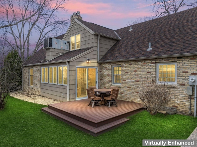 back of house at dusk with a chimney, roof with shingles, a wooden deck, outdoor dining space, and a yard