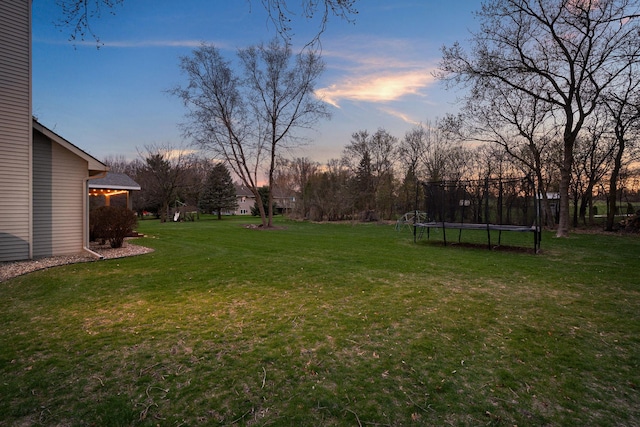 yard at dusk featuring a trampoline, a lawn, and view of wooded area