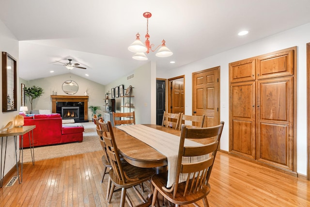 dining room with light wood finished floors, ceiling fan, a warm lit fireplace, vaulted ceiling, and a chandelier