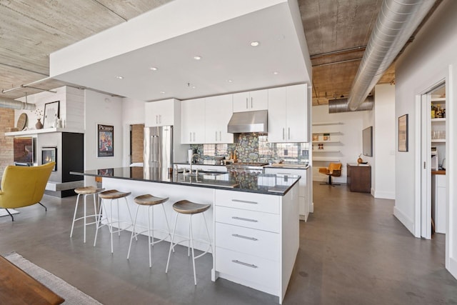 kitchen featuring under cabinet range hood, high end fridge, finished concrete floors, a sink, and decorative backsplash