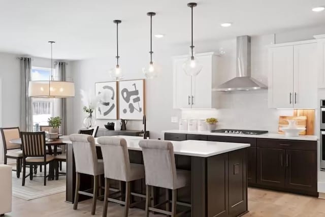 kitchen featuring wall chimney range hood, light countertops, an island with sink, white cabinets, and a kitchen bar