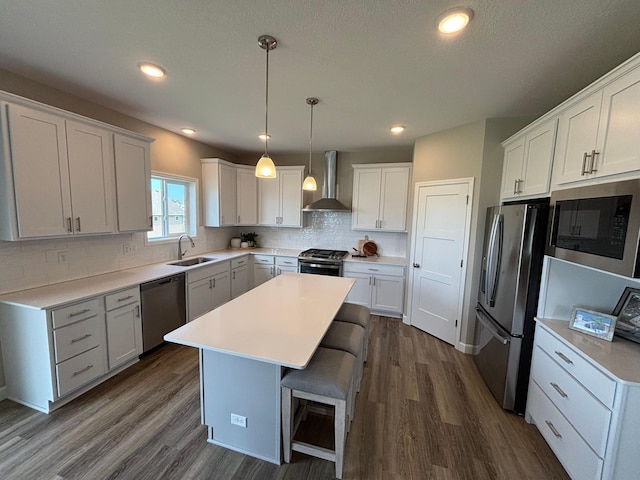kitchen featuring wall chimney range hood, appliances with stainless steel finishes, a sink, dark wood-type flooring, and a kitchen island