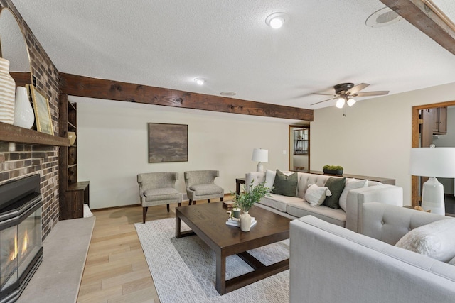 living room featuring ceiling fan, a textured ceiling, light wood finished floors, beam ceiling, and a brick fireplace