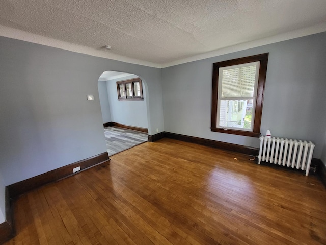 unfurnished room with radiator heating unit, arched walkways, hardwood / wood-style flooring, baseboards, and a textured ceiling