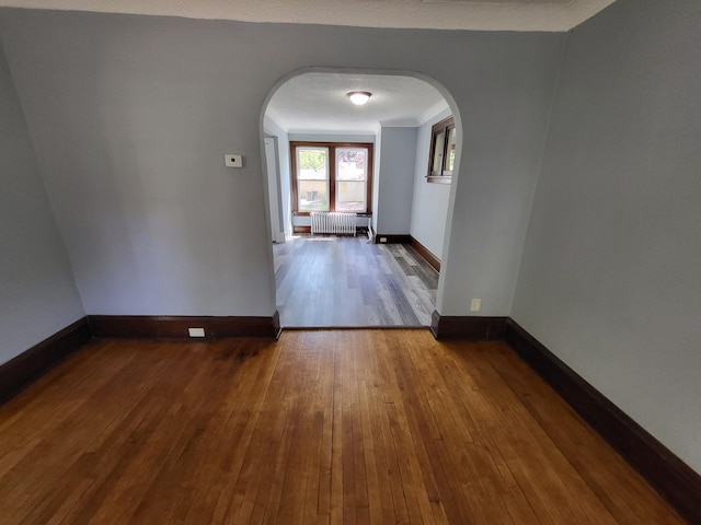 spare room featuring arched walkways, radiator heating unit, hardwood / wood-style flooring, and baseboards