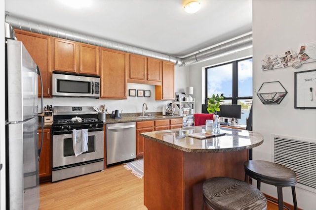 kitchen with appliances with stainless steel finishes, a kitchen breakfast bar, light wood-style flooring, dark stone countertops, and a sink