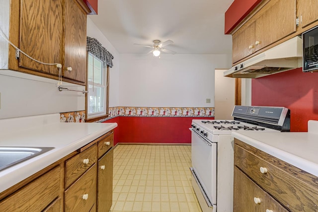 kitchen with white gas stove, ceiling fan, light countertops, brown cabinetry, and wainscoting